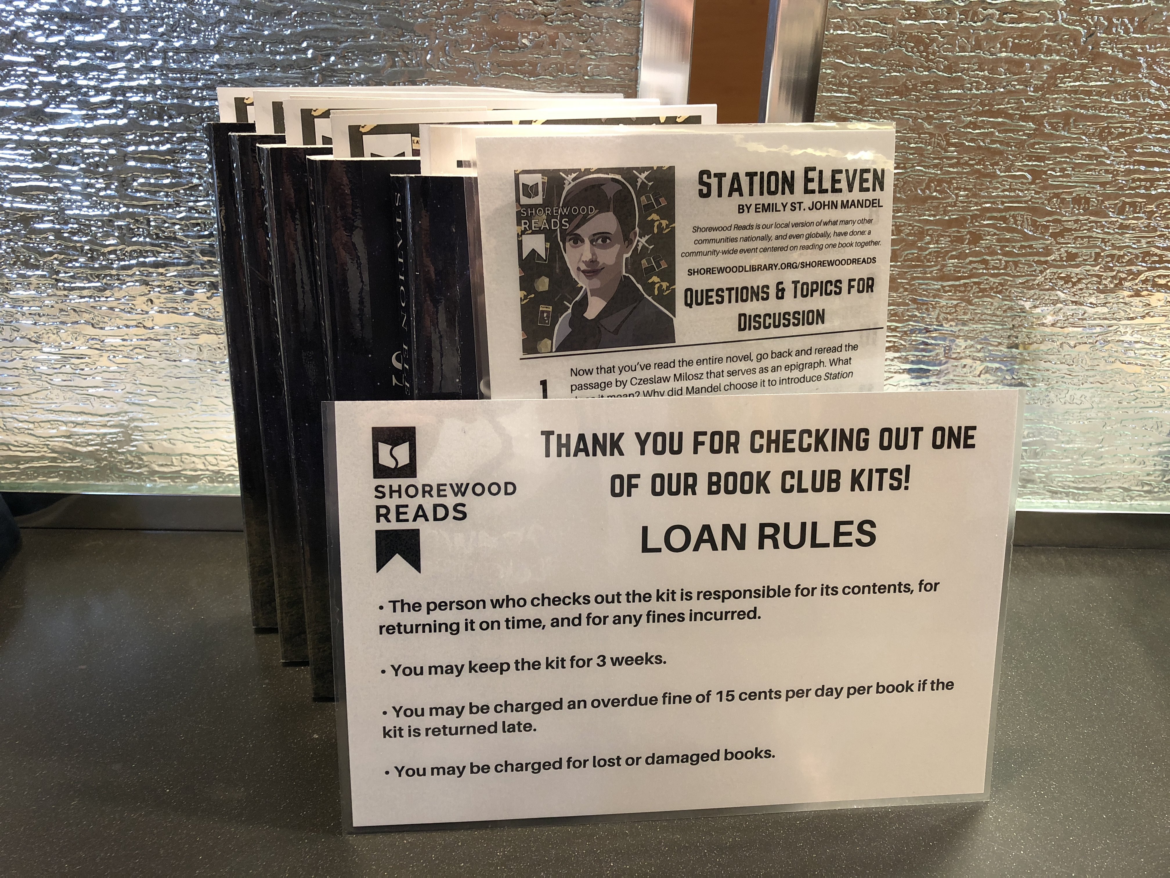 Copies of Station Eleven by a Shorewood Reads Loan Rules sign propped.